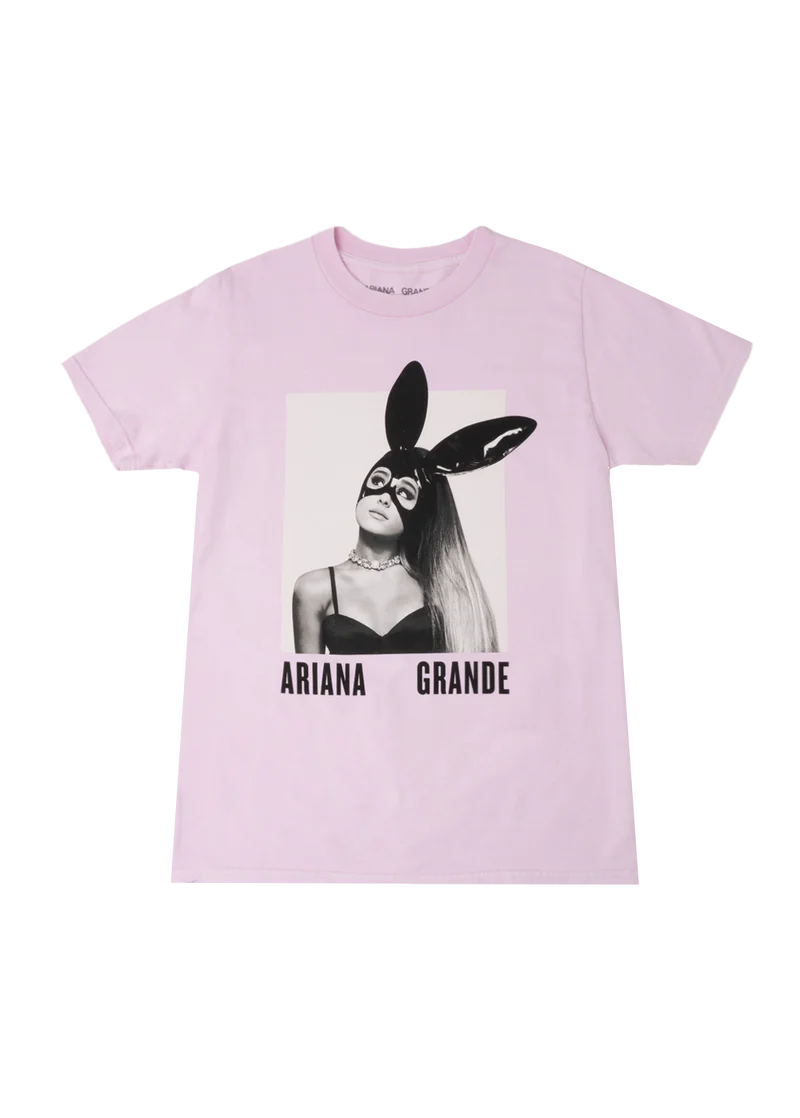 Light pink Ariana Grande t-shirt with a centered black-and-white graphic of Ariana wearing her iconic bunny mask from the Dangerous Woman album cover. The shirt features bold “Ariana Grande” text beneath the image, making it standout concert merch.
